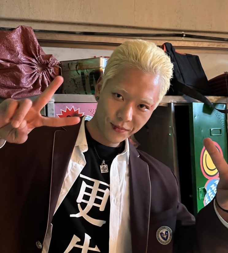 Joo Yeon-woo de cabelo platinado e uniforme escolar, fazendo sinal de V nos bastidores