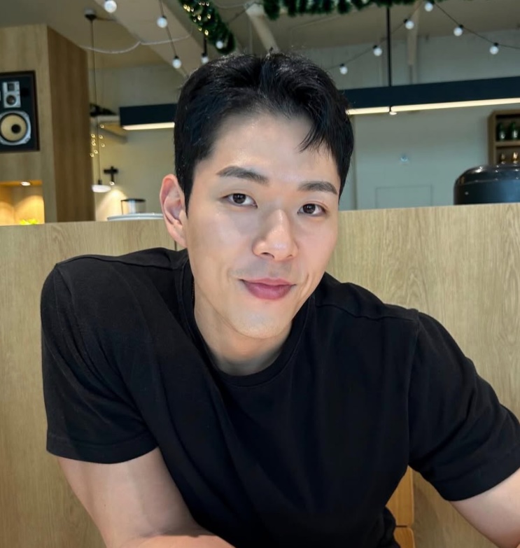 Joo Yeon-woo de camiseta preta, apoiado na mesa de uma cafeteria, sorrindo para a câmera