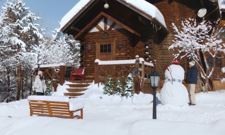 Pátio nevado diante da cabana, com um banco de madeira e um boneco de neve