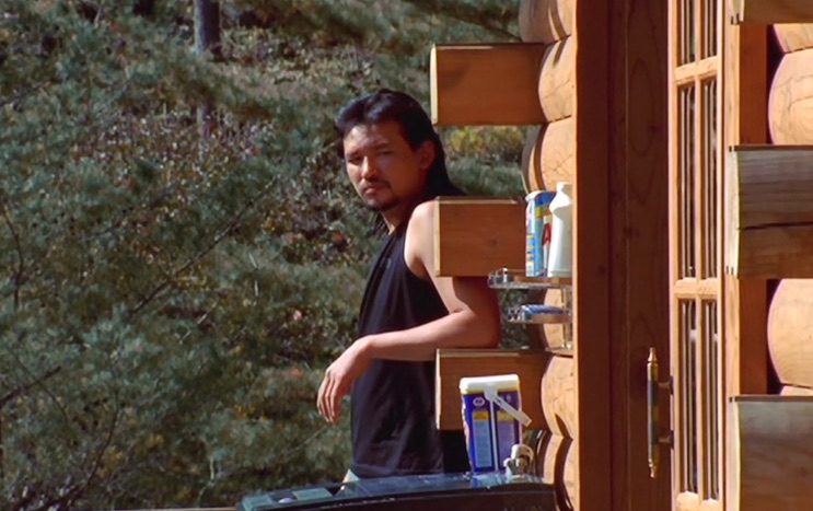 Homem de cabelo comprido olhando para trás na porta de uma cabana de troncos, em cena do filme Road Movie