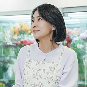 Bae Yoon-ok de avental florido, sorrindo dentro da floricultura
