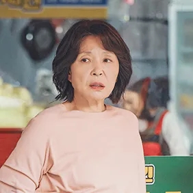 Ok Seon-ja de blusa rosa, em pé dentro da lanchonete
