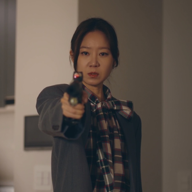Yu Bo-na de casaco cinza e lenço xadrez, apontando uma pistola dentro de casa