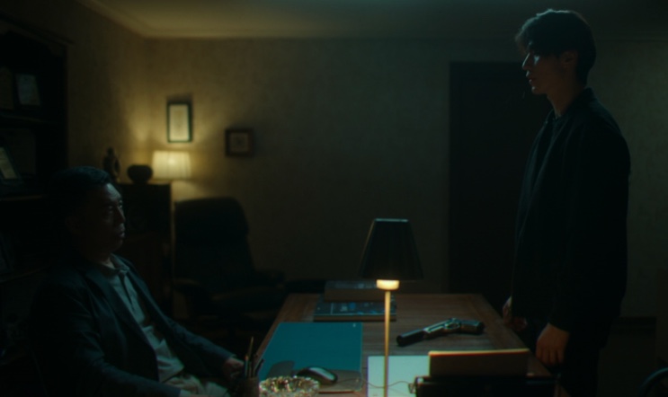 O Jin-man diante do chefe da Babylon, com uma arma sobre a mesa, em A Shop for Killers
