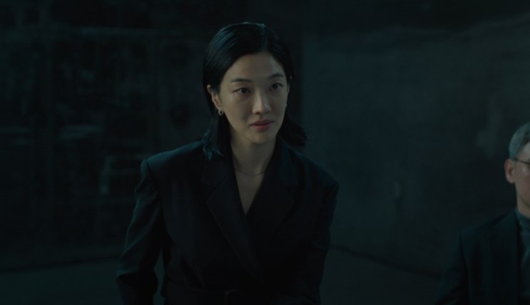 Kusanagi Jin de terno escuro, em pé num ambiente mal iluminado