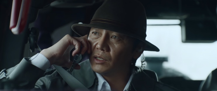 Kang Guk-cheol de chapéu, ao telefone no carro