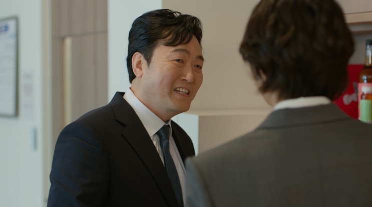 Bong Ji-uk de terno, sorrindo enquanto conversa com alguém de costas
