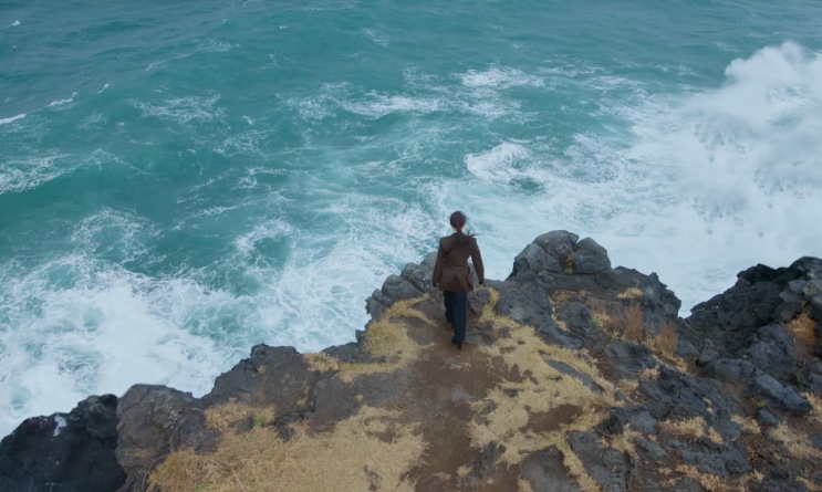 Yeo-ri de pé na ponta do penhasco, com o mar batendo nas rochas