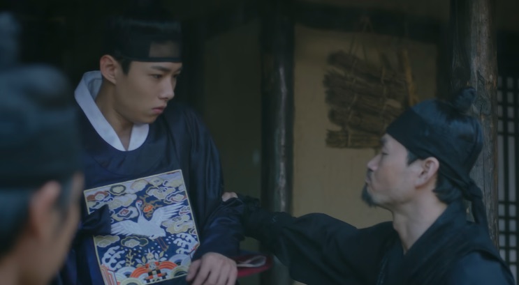Seo Ji-hoon sendo agarrado pela roupa por outro homem, segurando uma caixa