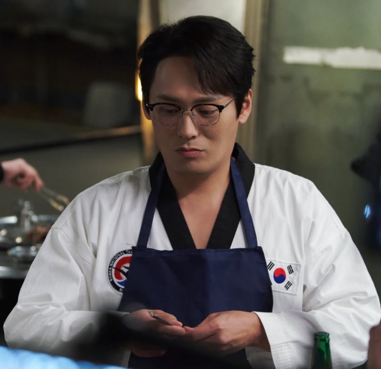 Choi Dae-hoon de uniforme de taekwondo (dobok) e avental, como Sung Han-soo