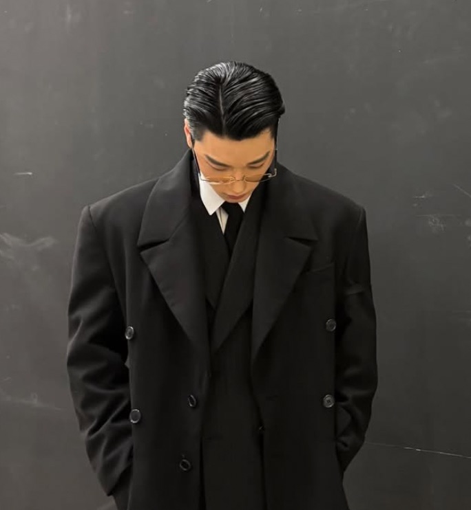 Choi San de sobretudo preto e cabelo penteado para trás, olhando para baixo