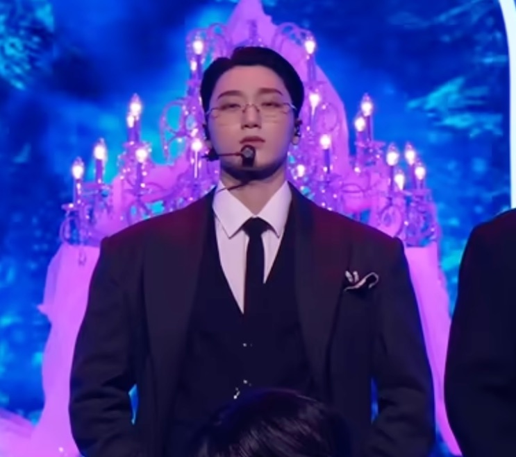Choi San no palco de terno de três peças, com um lustre iluminado atrás