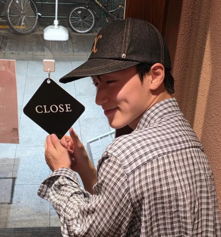 Choi San de boné e camisa xadrez, sorrindo ao lado de uma plaquinha 'CLOSE'