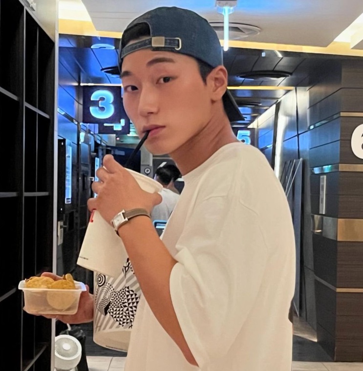 Choi San de boné virado ao contrário, tomando uma bebida e segurando um pote de batata