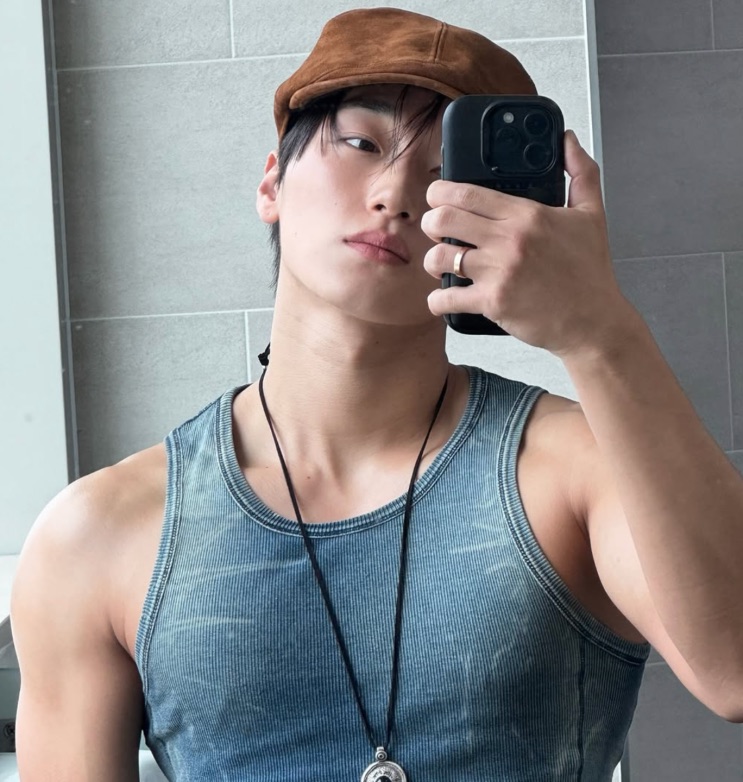 Selfie de Choi San no espelho, de regata e boné, mostrando os braços