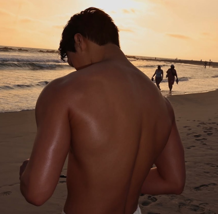 Choi San atual de costas na praia ao pôr do sol, ombros e costas trabalhados