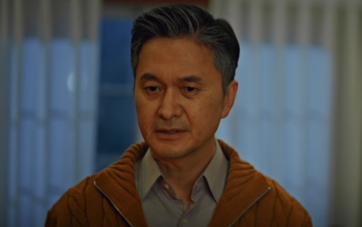 O presidente Jin Myung-chul de suéter marrom, de pé num corredor