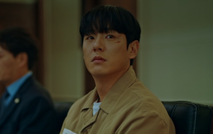 Jin Yi-soo sentado, de jaqueta bege, com expressão dura