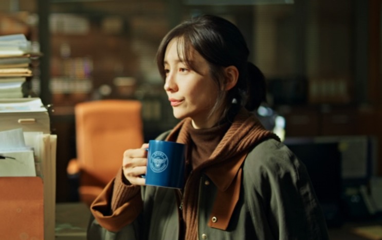 Lee Kang-hyun de casaco, segurando uma caneca azul na delegacia
