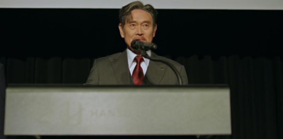 Choi Jung-hoon discursando ao microfone num púlpito com o nome Hansu