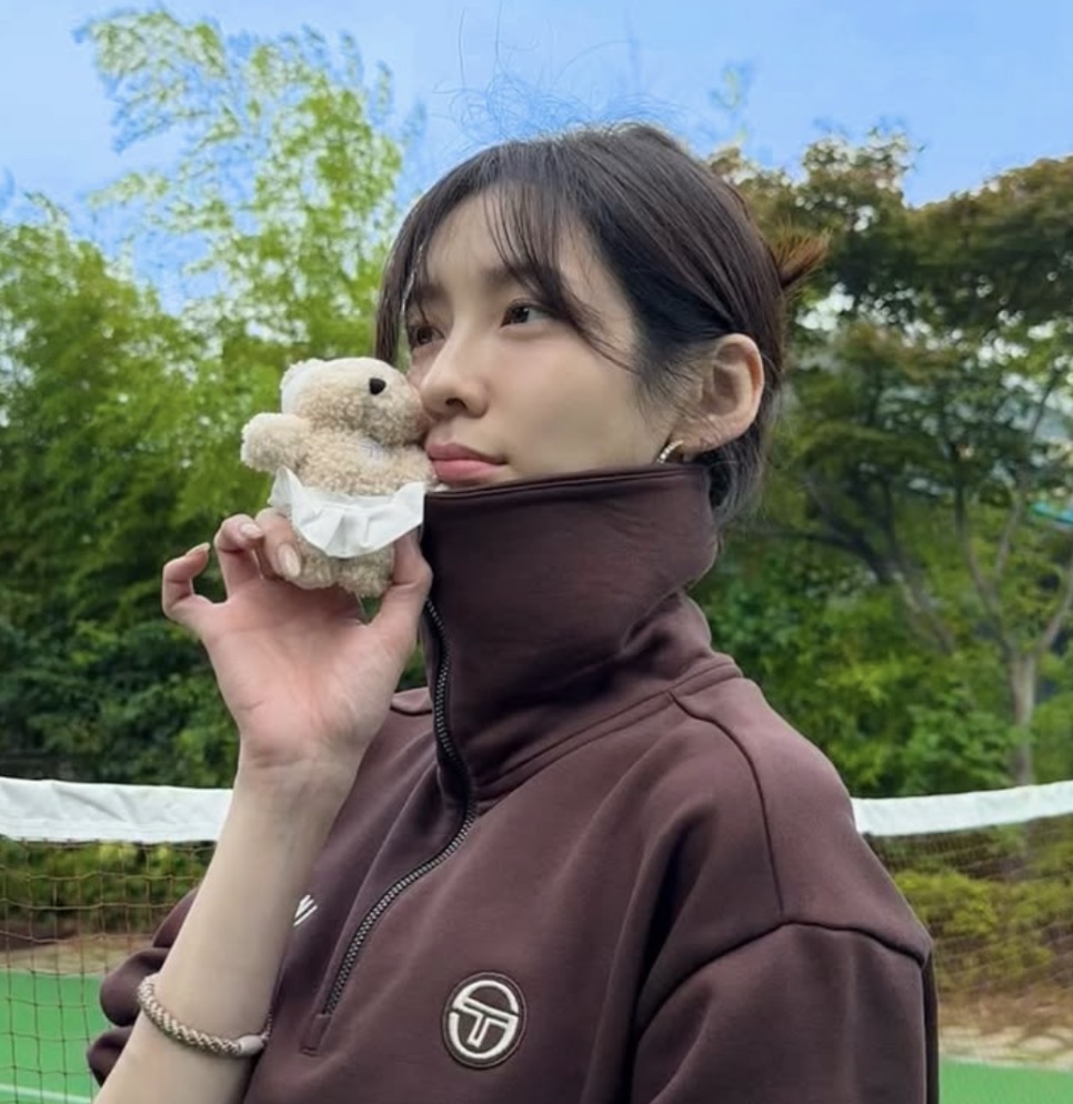 Park Ji-hyun de casaco marrom numa quadra de tênis, segurando um ursinho de pelúcia
