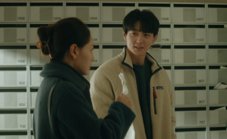 Ji-su e Woo-jae conversando de pé no corredor das caixas de correio