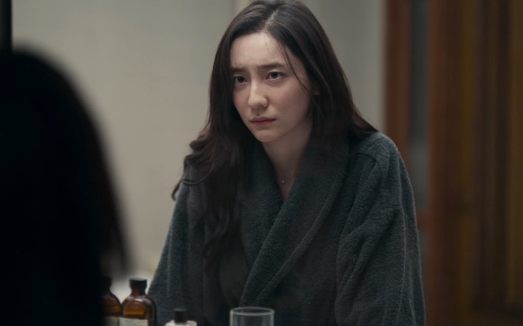Park Ji-hyun de roupão cinza, diante do espelho, com vidros sobre a bancada