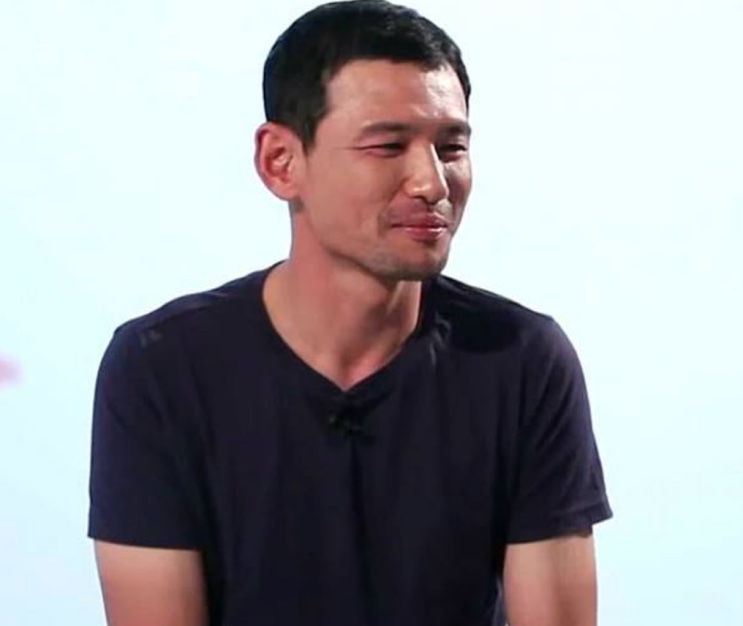 Hwang Jung-min de camiseta escura, sorrindo de lado, sobre fundo claro