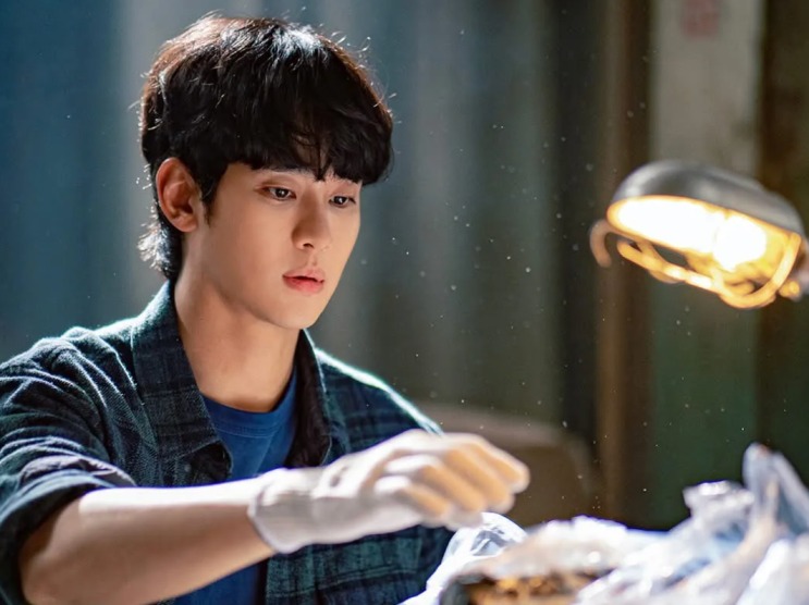 Cena de Knock-Off: Kim Soo-hyun de camisa xadrez, mexendo em algo sobre uma mesa