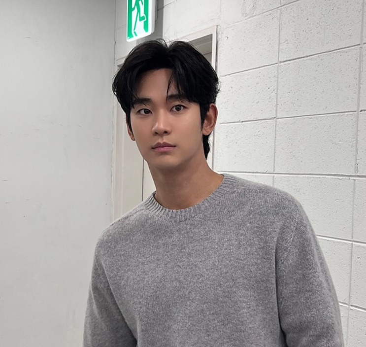 Kim Soo-hyun de suéter cinza, encostado numa parede branca