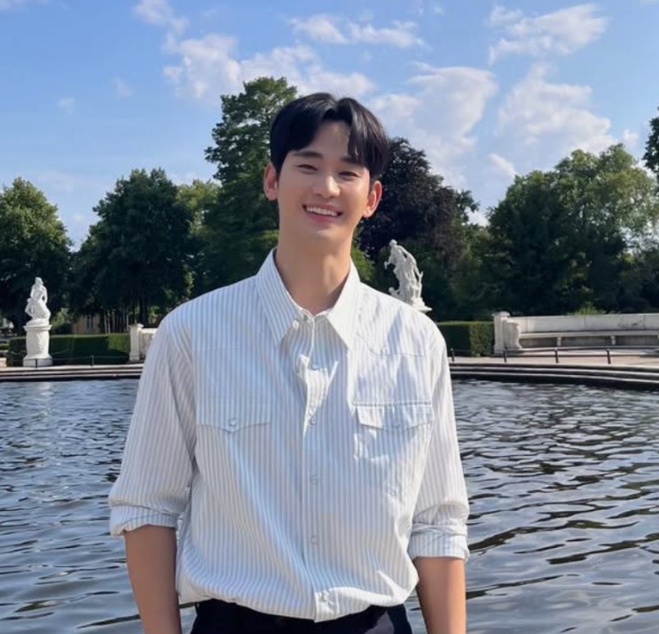 Kim Soo-hyun de camisa listrada, sorrindo, diante de um lago