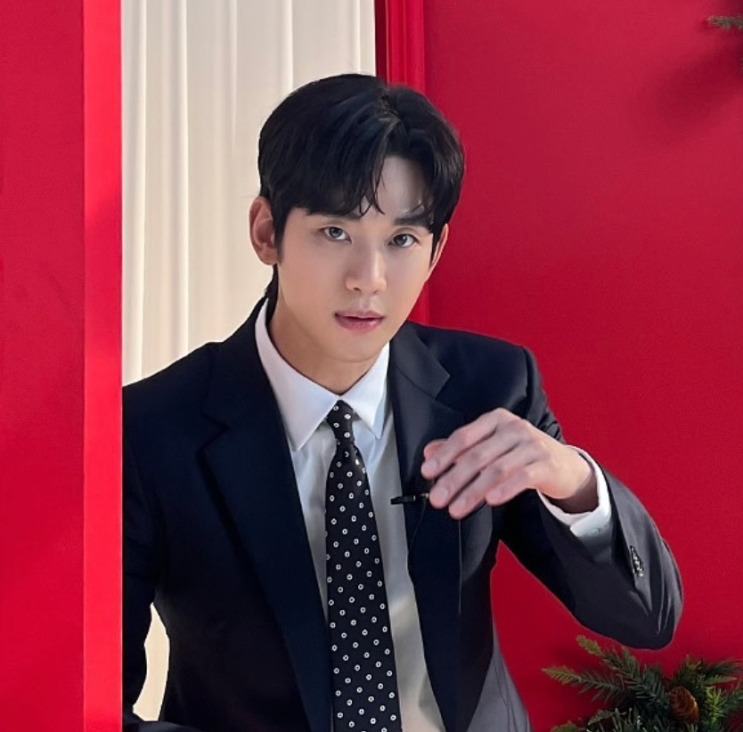 Kim Soo-hyun de terno escuro e gravata de bolinhas, diante de um fundo vermelho