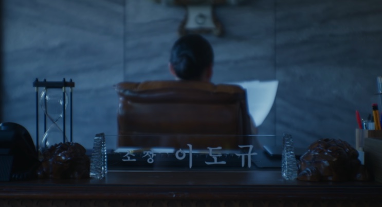 A mesa do diretor da agência Baekho, com a placa de nome do Lee Do-gyu e a cadeira virada de costas