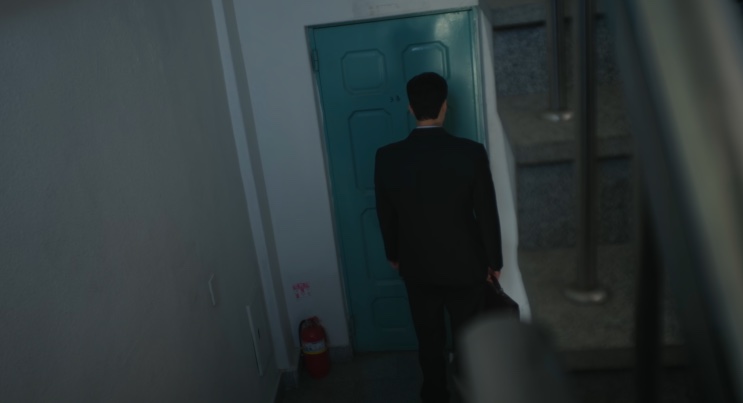 Kim de terno, de costas, parado diante da porta azul da agência Baekho