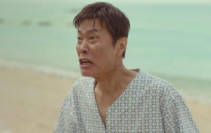 Ri Eung-ryeong, com cicatrizes no rosto e roupa de hospital, numa praia