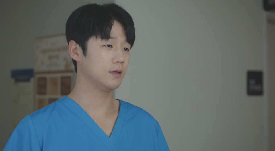 Jung Joon-won de pijama cirúrgico azul, num corredor de hospital