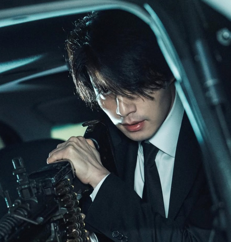 Lee Dong-wook de terno, com uma arma, dentro de um carro