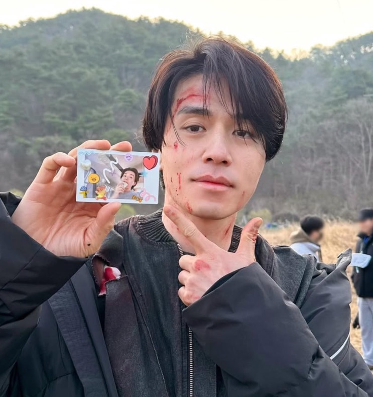Lee Dong-wook nos bastidores, com o rosto sujo de sangue cênico, segurando um photocard