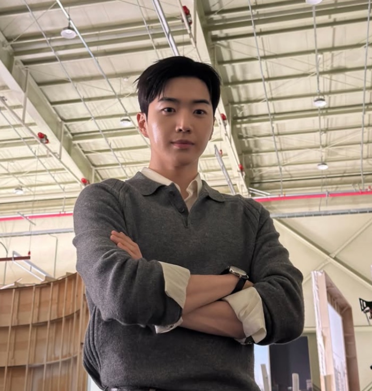 Kang Ha-gi de suéter cinza e braços cruzados, num escritório amplo