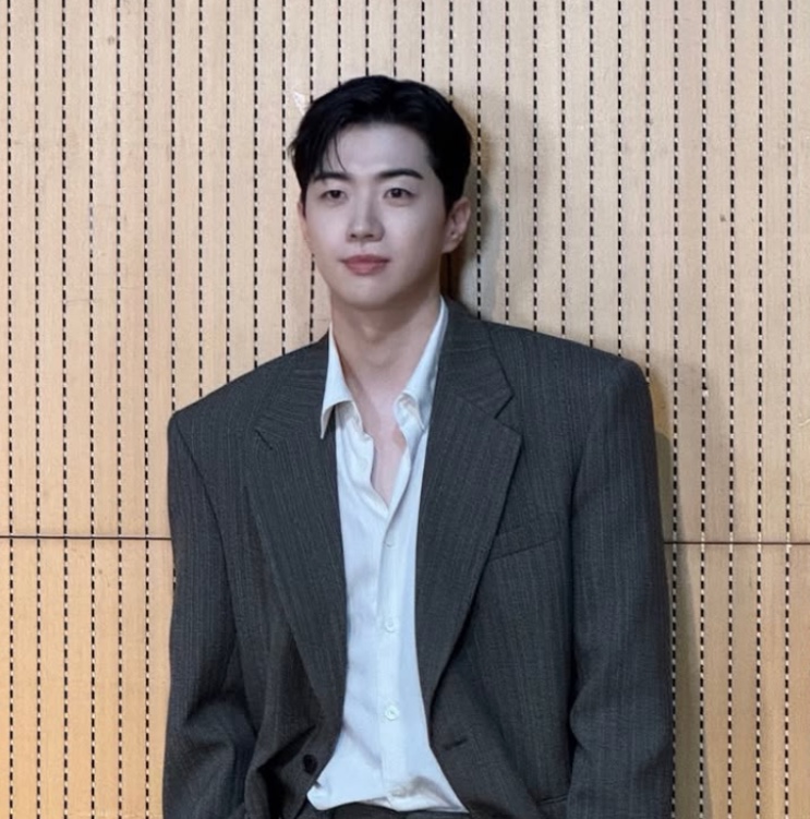 Kang Hoon de blazer cinza e camisa branca aberta, encostado numa parede de madeira