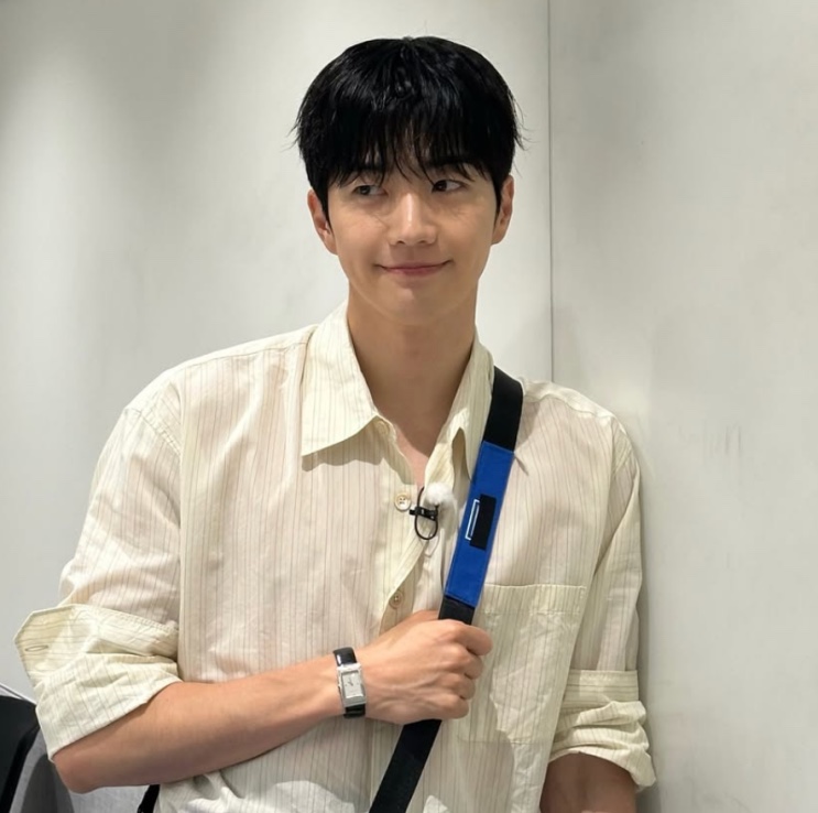 Kang Hoon sorrindo de canto, de camisa listrada, segurando a alça de uma bolsa