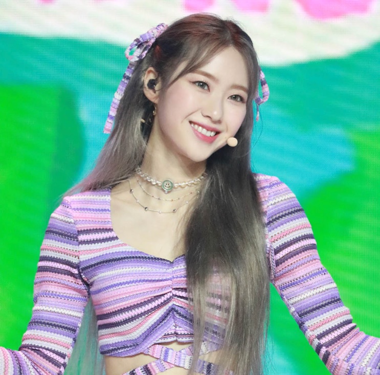 Jiho no palco, de figurino lilás e microfone headset, sorrindo