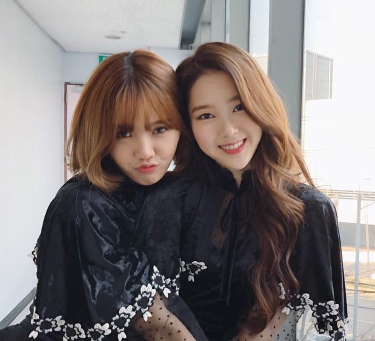 Jiho ao lado de outra integrante do grupo, as duas de vestido preto nos bastidores