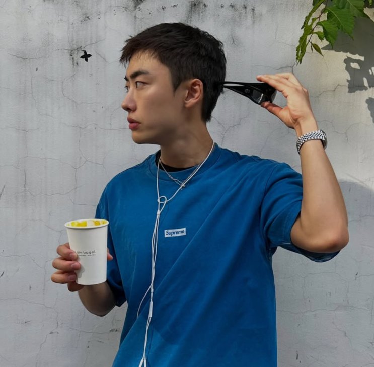 Lee Won-jung de camiseta azul, segurando um copo, passando a máquina no próprio cabelo