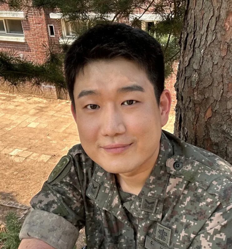 Nam Tae-woo de uniforme camuflado, sentado à sombra de um pinheiro