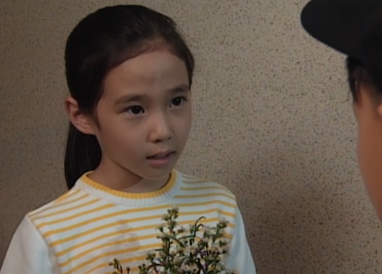 Su-ji de blusa listrada segurando flores, de perfil, conversando com alguém