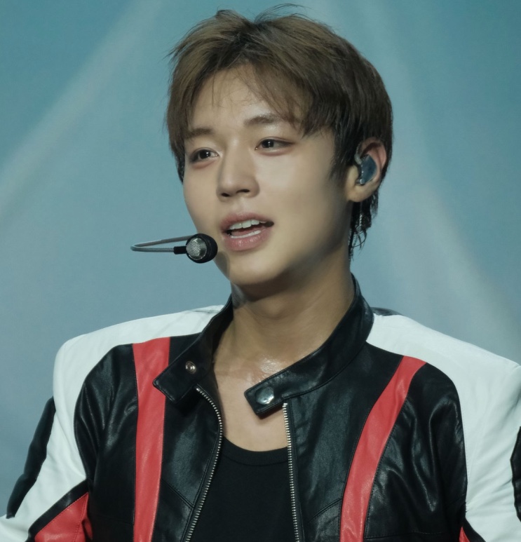 Park Ji-hoon cantando no palco, de jaqueta preta e ponto in-ear na orelha