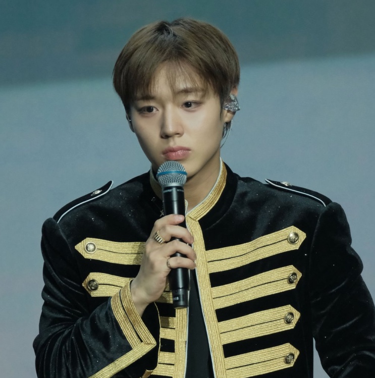 Park Ji-hoon no palco, ao microfone, com jaqueta preta de detalhes dourados