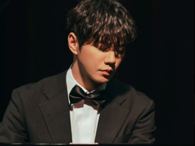 Lee Jun-young de smoking e gravata-borboleta, sentado no escuro