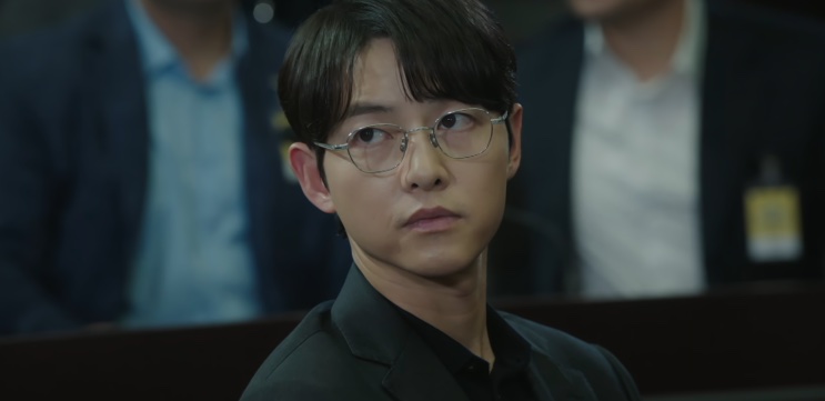 Yoon Hyun-woo de óculos, olhando para a frente em um ambiente escuro